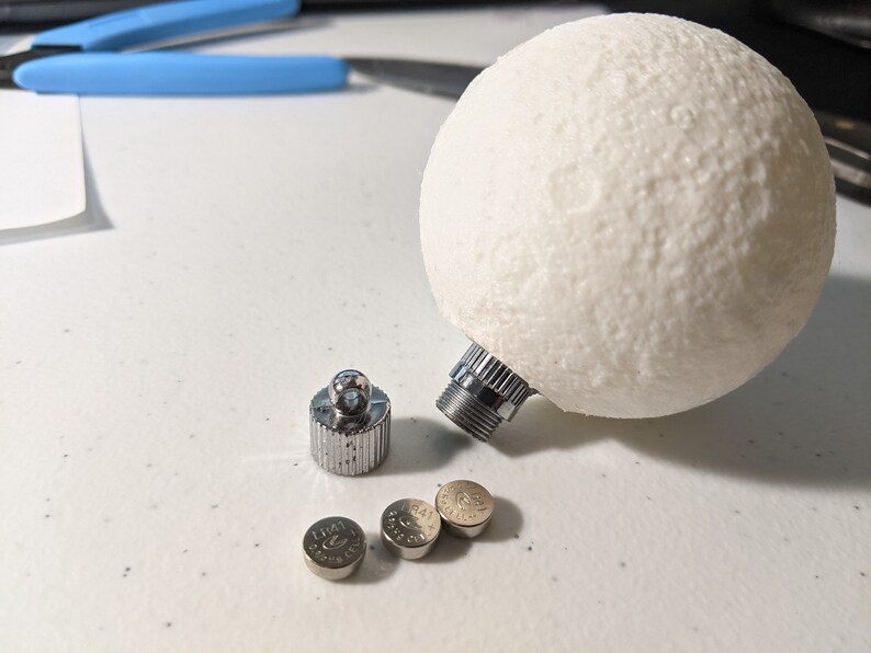 May include: A white, textured sphere with a silver metal base, resembling the moon. Beside it are a silver metal cap and three small, round batteries. The items are on a white surface.