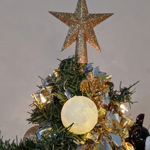 May include: A small Christmas tree decorated with ornaments and lights. A gold glitter star tree topper sits at the top. The tree has a white, textured ball ornament, a red ornament, and gold tinsel.