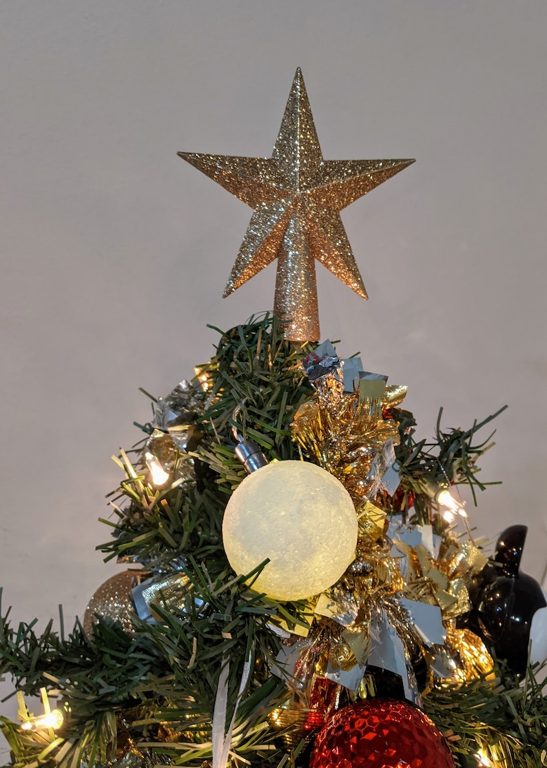 May include: A small Christmas tree decorated with ornaments and lights. A gold glitter star tree topper sits at the top. The tree has a white, textured ball ornament, a red ornament, and gold tinsel.