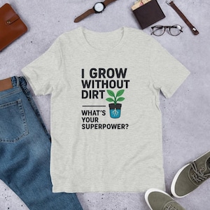 Puede incluir: Camiseta gris claro con el texto "I GROW WITHOUT DIRT" sobre un gráfico de una planta en una maceta, y el texto "WHAT'S YOUR SUPERPOWER?" debajo. La camiseta está hecha de un material suave.