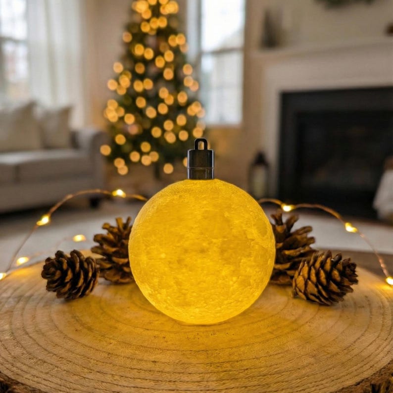 Glowing yellow 3D-printed mini moon ornament resting on a rustic wood slice. Surrounded by pinecones and soft fairy lights with a blurred lit Christmas tree in the background. Shows the textured lunar surface.
