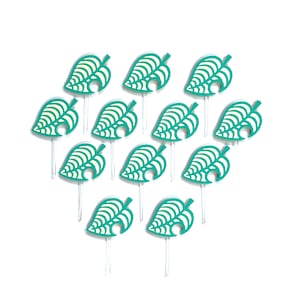 May include: A collection of teal leaf-shaped toppers with white detailing, arranged on clear sticks. The toppers are arranged in a triangular pattern against a white background. The design is reminiscent of a popular video game.