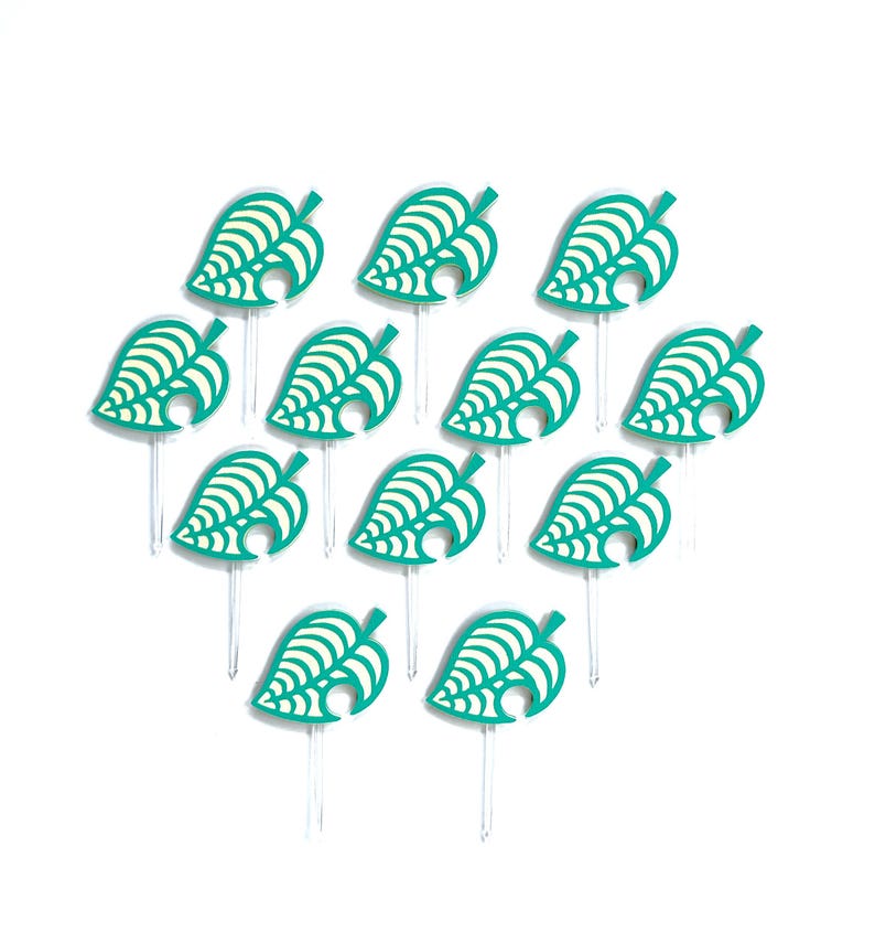 May include: A collection of teal leaf-shaped toppers with white detailing, arranged on clear sticks. The toppers are arranged in a triangular pattern against a white background. The design is reminiscent of a popular video game.