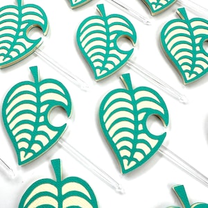May include: Close-up of several teal and cream-colored leaf-shaped toppers with clear plastic stems. The leaves have a detailed vein design and a distinctive curved edge. The toppers are arranged on a white surface, suggesting a party or craft theme.
