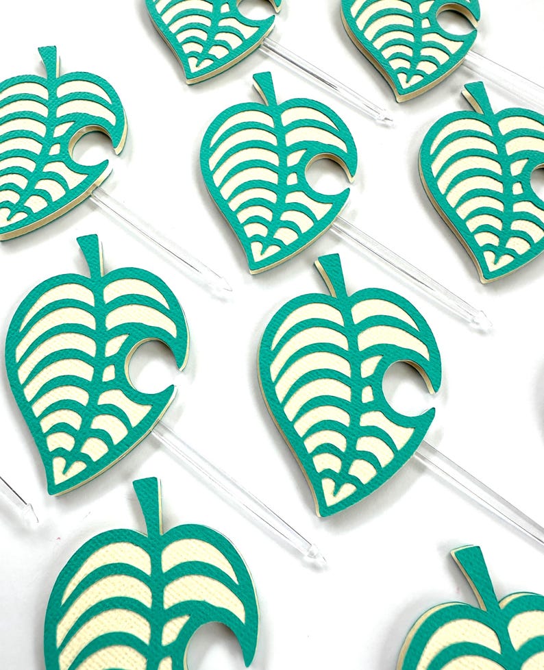 May include: Close-up of several teal and cream-colored leaf-shaped toppers with clear plastic stems. The leaves have a detailed vein design and a distinctive curved edge. The toppers are arranged on a white surface, suggesting a party or craft theme.