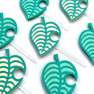 May include: Close-up of several teal and cream-colored leaf-shaped toppers with clear plastic sticks. The leaves have a layered design with a cutout on one side. The toppers are arranged on a white surface.