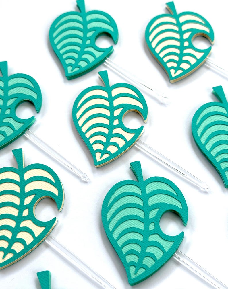 May include: Close-up of several teal and cream-colored leaf-shaped toppers with clear plastic sticks. The leaves have a layered design with a cutout on one side. The toppers are arranged on a white surface.