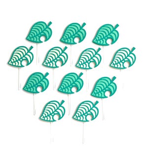 May include: A collection of teal and white leaf-shaped cupcake toppers. Each topper features a detailed leaf design with a stem and a small hole. The toppers are arranged on clear plastic sticks against a white background.