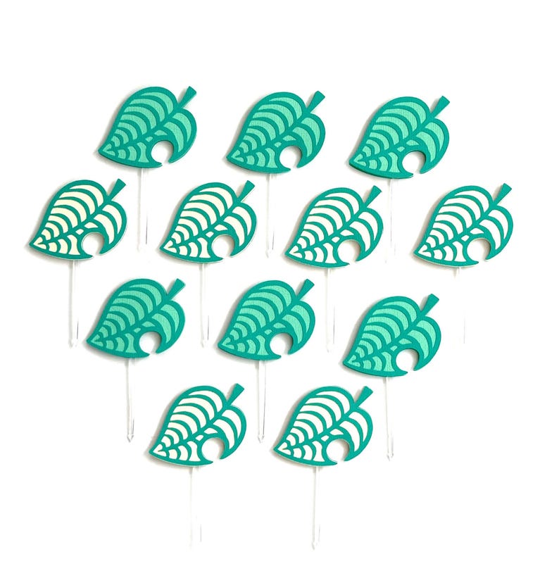May include: A collection of teal and white leaf-shaped cupcake toppers. Each topper features a detailed leaf design with a stem and a small hole. The toppers are arranged on clear plastic sticks against a white background.
