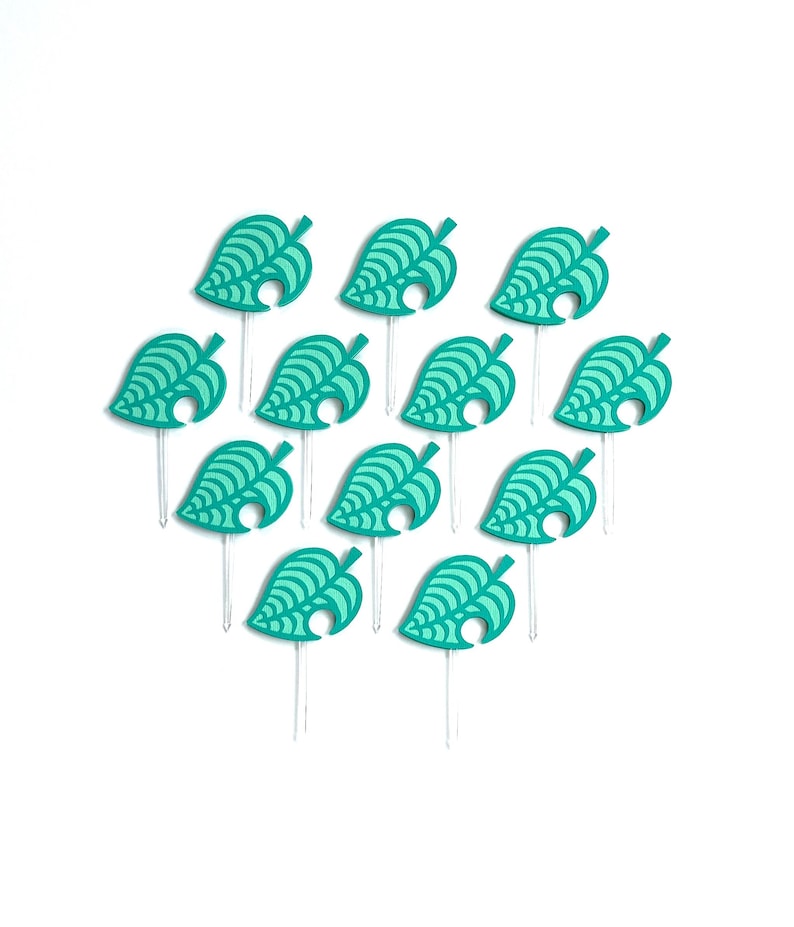 May include: A collection of teal leaf-shaped cupcake toppers. Each topper features a detailed leaf design and is attached to a clear plastic stick. The toppers are arranged on a white background.