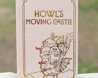 May include: Hardback book with the title "Howl's Moving Castle" in gold lettering. The cover has a gold illustration of the castle and a decorative gold border. A white ribbon bookmark is visible.