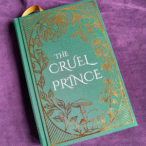 May include: A hardcover book titled "The Cruel Prince" with a teal cover and gold foil floral and mushroom illustrations. The book has a gold ribbon bookmark and is set against a purple background.