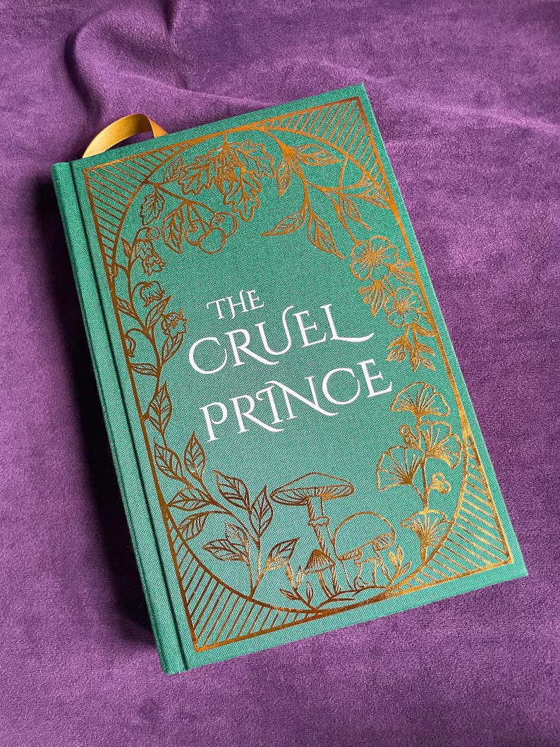May include: A hardcover book titled "The Cruel Prince" with a teal cover and gold foil floral and mushroom illustrations. The book has a gold ribbon bookmark and is set against a purple background.