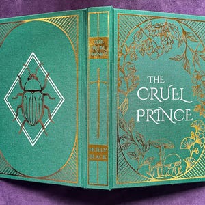 May include: Hardcover book titled "The Cruel Prince" by Holly Black. The book has a teal cover with gold foil accents, including a beetle illustration and floral designs. The spine features a gold sword and the title.