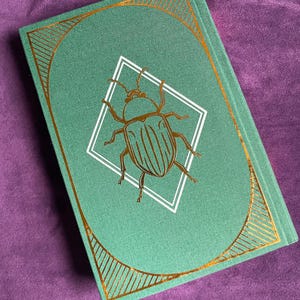 May include: A hardcover journal with a teal cover featuring a gold-colored beetle illustration inside a white diamond. The cover has a gold geometric border and a gold ribbon bookmark. The journal is closed and resting on a purple surface.