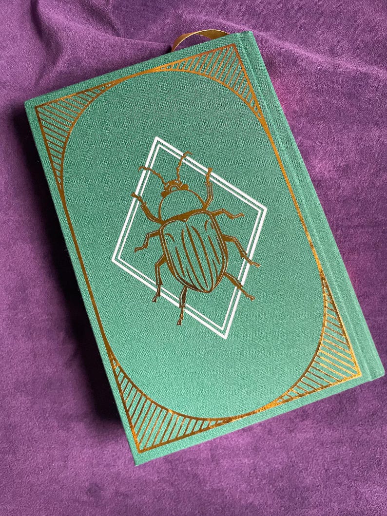 May include: A hardcover journal with a teal cover featuring a gold-colored beetle illustration inside a white diamond. The cover has a gold geometric border and a gold ribbon bookmark. The journal is closed and resting on a purple surface.