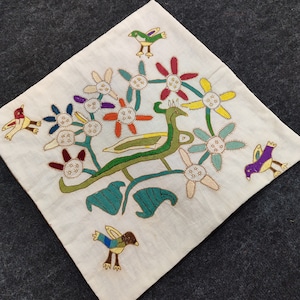 May include: Cream-colored square textile with embroidered design. Features a central green and yellow creature with a long neck, surrounded by colorful flowers and several embroidered birds in various colors. The textile is placed on a dark gray surface.