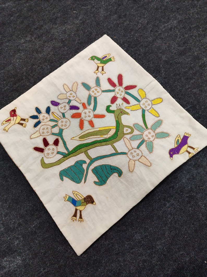 May include: Cream-colored square textile with embroidered design. Features a central green and yellow creature with a long neck, surrounded by colorful flowers and several embroidered birds in various colors. The textile is placed on a dark gray surface.