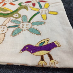 May include: Cream-colored textile with colorful embroidered designs. Features a purple bird with yellow accents, green serpent-like creature, and various flowers in red, yellow, and blue. The embroidery has a detailed, handmade appearance.
