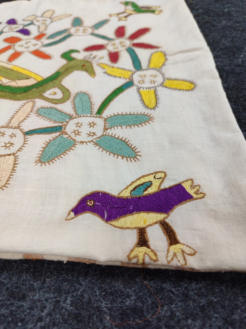 May include: Cream-colored textile with colorful embroidered designs. Features a purple bird with yellow accents, green serpent-like creature, and various flowers in red, yellow, and blue. The embroidery has a detailed, handmade appearance.