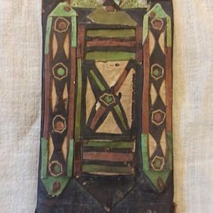 May include: A small, square, leather pouch with intricate geometric designs in brown, green, and white. The pouch is decorated with stitched leather strips and has a flap closure.