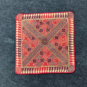 May include: A red, cream, and purple embroidered fabric square with a geometric pattern. The fabric has a pink border.