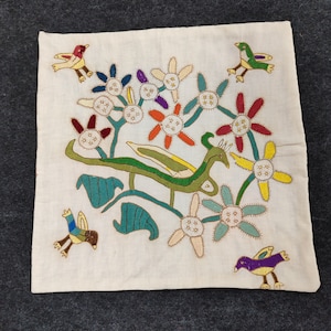 May include: Cream-colored square textile with embroidered design. Features colorful flowers, green leaves, and stylized birds in flight. The embroidery uses various colors including red, yellow, blue, and purple.