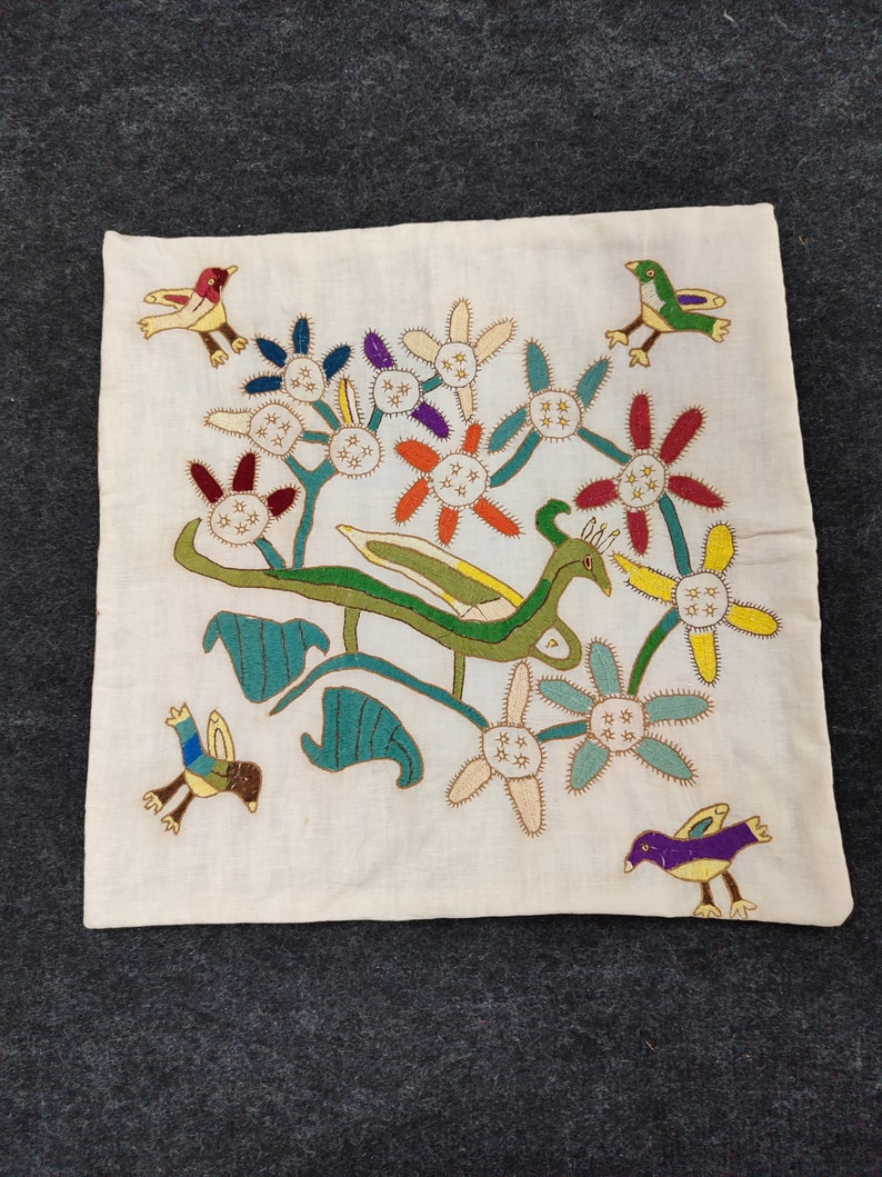 May include: Cream-colored square textile with embroidered design. Features colorful flowers, green leaves, and stylized birds in flight. The embroidery uses various colors including red, yellow, blue, and purple.