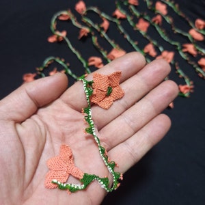 May include: Close-up of handmade crochet floral trim. The trim features peach-colored flowers with green centers and stems, and a white and green border. The delicate design is displayed against a dark background.