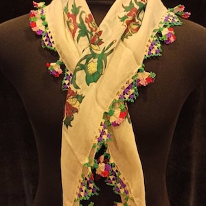 May include: A white cotton scarf with a floral print and a crocheted border in shades of green, purple, and pink.