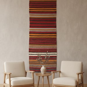 May include: A long, rectangular, striped rug in shades of red, orange, and brown hangs on a neutral wall. Two cream-colored armchairs and a small wooden side table with a vase of dried flowers are in front of the rug.