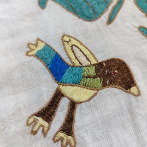 May include: Embroidered textile featuring a colorful bird and stylized leaves. The bird has blue, green, and brown sections, with yellow accents. The leaves are teal with brown outlines, all on a white background.