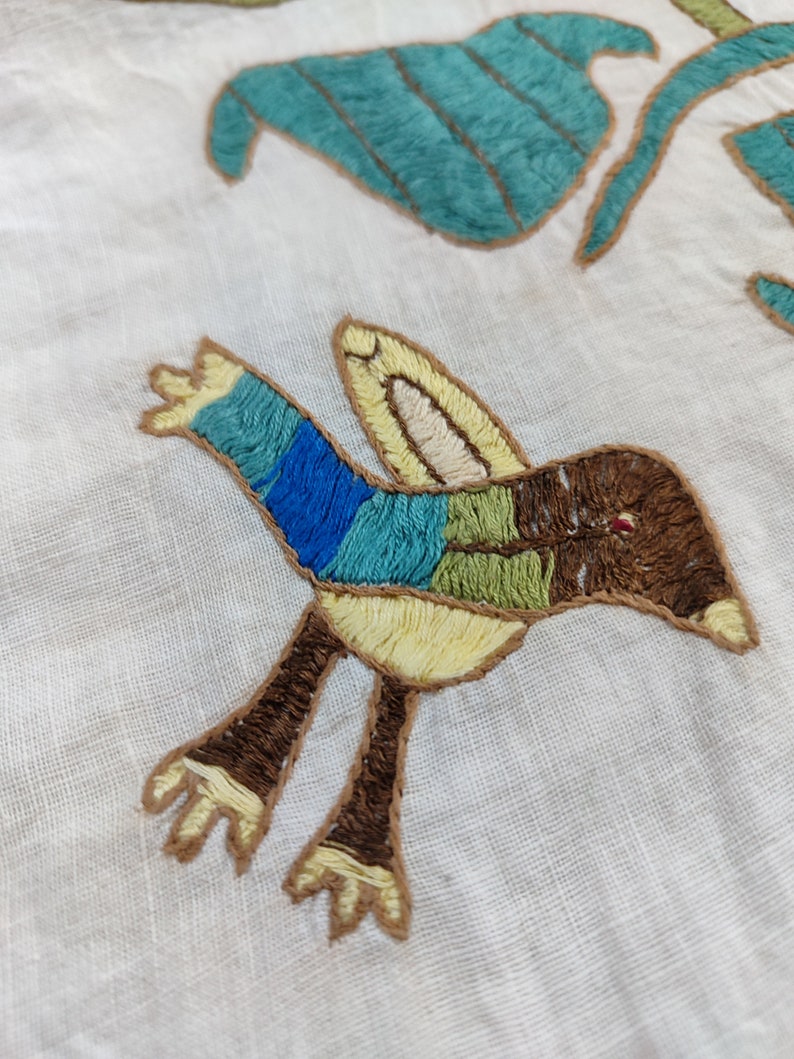 May include: Embroidered textile featuring a colorful bird and stylized leaves. The bird has blue, green, and brown sections, with yellow accents. The leaves are teal with brown outlines, all on a white background.