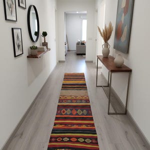 May include: A hallway with a colorful, patterned runner rug. The rug features a geometric design with red, orange, yellow, and black stripes. The walls are white, with framed artwork and a mirror. A wooden console table with vases is on the right.