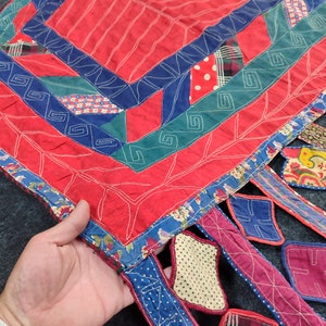 May include: A red, blue, and green patchwork quilt with a square design in the center and fringed edges. The quilt is made of various fabrics, including floral prints, polka dots, and plaid.
