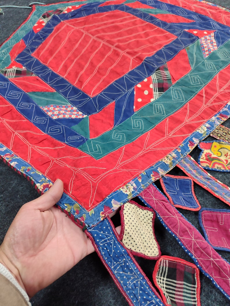 May include: A red, blue, and green patchwork quilt with a square design in the center and fringed edges. The quilt is made of various fabrics, including floral prints, polka dots, and plaid.