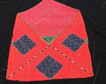 May include: A red quilted fabric pouch with a geometric pattern. The pouch is made of red fabric with black and green accents. The pouch is folded in half and has a triangular shape.