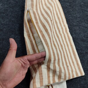 May include: A striped, beige and brown fabric item with a zipper and a pocket. The item has a vertical stripe pattern. The pocket is open, revealing a mesh lining. The item is on a dark gray surface.