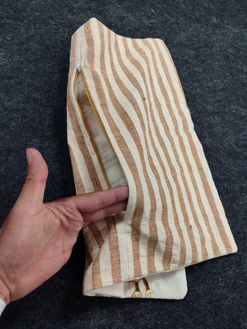 May include: A striped, beige and brown fabric item with a zipper and a pocket. The item has a vertical stripe pattern. The pocket is open, revealing a mesh lining. The item is on a dark gray surface.