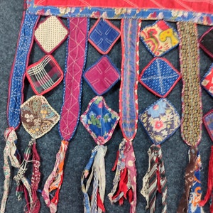 May include: A colorful patchwork textile with a fringe of fabric scraps. The textile is made of various fabrics in shades of red, blue, green, and white. The fringe is made of strips of fabric in various colors and patterns.