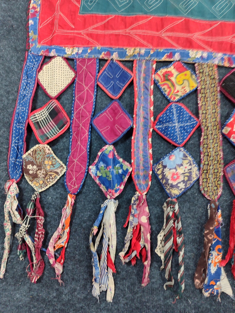 May include: A colorful patchwork textile with a fringe of fabric scraps. The textile is made of various fabrics in shades of red, blue, green, and white. The fringe is made of strips of fabric in various colors and patterns.