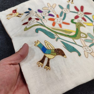 May include: Cream-colored embroidered textile featuring colorful birds and floral designs. The embroidery includes birds with blue, green, and brown feathers, and flowers with orange, red, and purple petals. The textile is held in a hand.