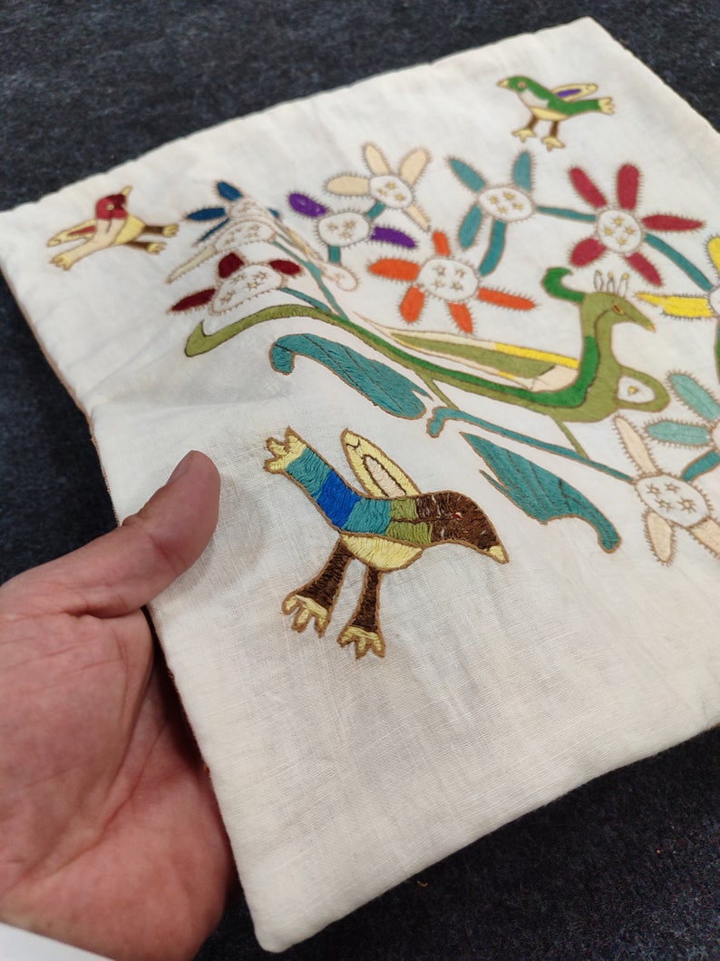 May include: Cream-colored embroidered textile featuring colorful birds and floral designs. The embroidery includes birds with blue, green, and brown feathers, and flowers with orange, red, and purple petals. The textile is held in a hand.