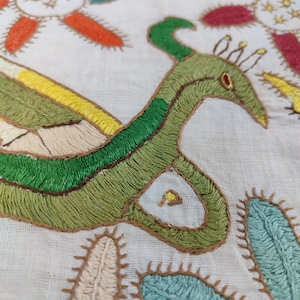 May include: Close-up of a white textile with colorful embroidery. The design features a green bird-like figure with a long neck and a red eye, surrounded by floral and cactus-like motifs in shades of orange, yellow, blue, and red.