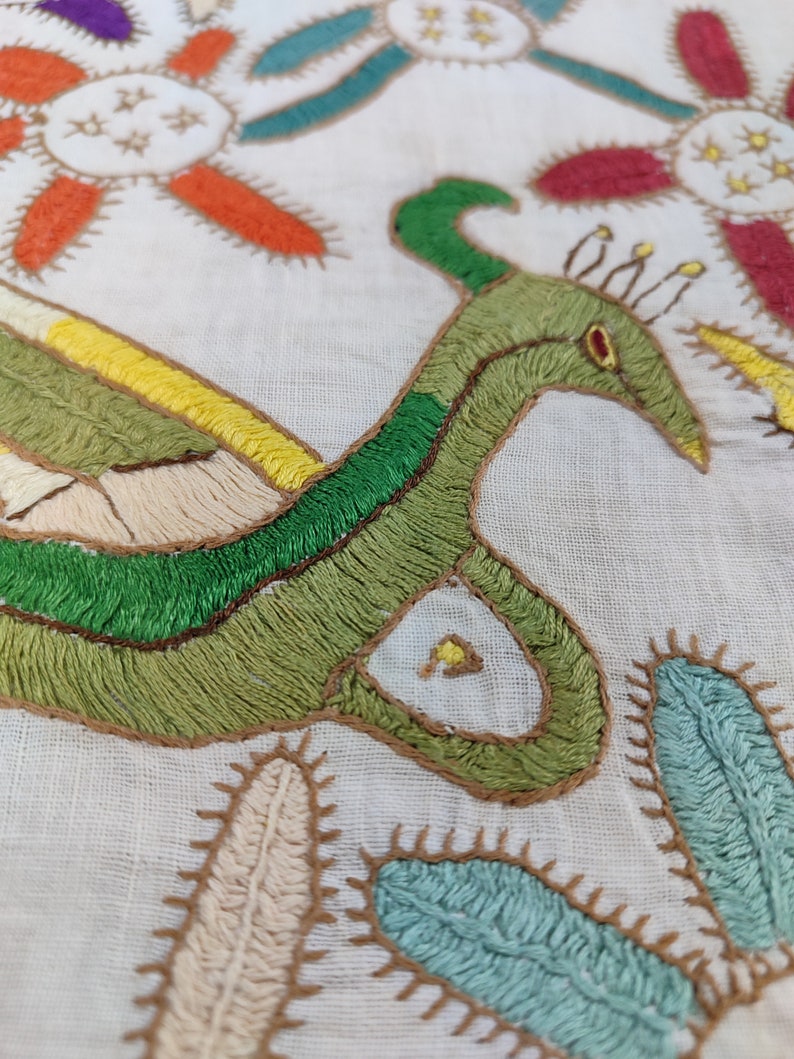 May include: Close-up of a white textile with colorful embroidery. The design features a green bird-like figure with a long neck and a red eye, surrounded by floral and cactus-like motifs in shades of orange, yellow, blue, and red.