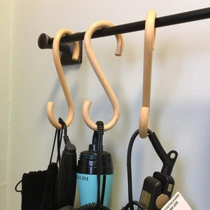 Pode incluir: Três ganchos de madeira pendurados em uma barra de metal preta. Os ganchos são em forma de 'S' e são usados para pendurar vários itens, incluindo uma ferramenta de modelagem de cabelo azul e preta, uma ferramenta de modelagem de cabelo preta e uma ferramenta de modelagem de cabelo preta e prateada.