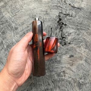 May include: A cylindrical handle with a dark brown and black wood grain pattern featuring a vibrant red swirl accent. The handle has a metallic silver threaded top fitting and is accompanied by a separate, matching red and black wood cap.
