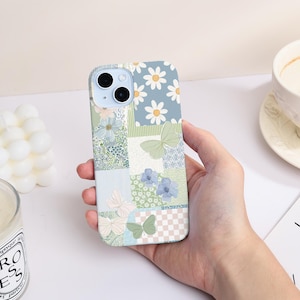 Op de afbeelding: Een telefoonhoesje met bloemenpatroon en patchwork-ontwerp met madeliefjes, vlinders en botanische elementen in blauw, groen en wit. De hoes wordt in een hand gehouden en toont het ontwerp.