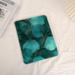 Puede incluir: Una funda para tableta de color verde azulado y dorado con un diseño de hojas de eucalipto en acuarela. La funda tiene forma rectangular redondeada y un patrón de hojas doradas. Un lápiz óptico blanco está al lado de la funda. La funda está sobre una superficie blanca.