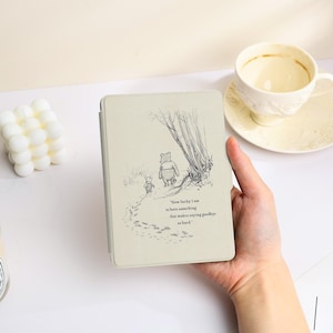 May include: A light beige e-reader cover featuring a black ink illustration of Winnie the Pooh and Piglet walking away. The text reads, "How lucky I am to have something that makes saying goodbye so hard." The cover has a smooth, matte finish.